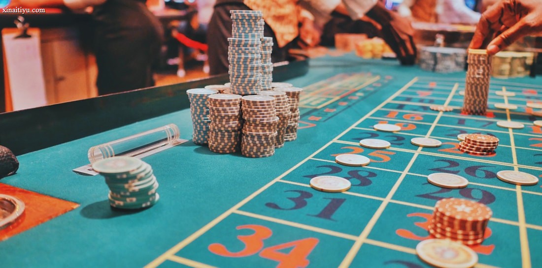 A casino scene with a roulette table features prominently, covered in green felt with colorful numbers and betting squares. Several stacks of poker chips are placed on different numbers, and the hands of several people are visible as they interact with the table. In the background, there are slot machines and more people, creating a vibrant and busy atmosphere.
