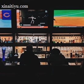 A bar counter with several people sitting in front of it is shown. Above the counter, there are multiple television screens displaying sports broadcasts, including football and baseball. Behind the counter, various bottles of alcohol are neatly arranged.
