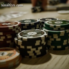 Stacks of poker chips are arranged on a wooden table, with various denominations and colors such as red, green, and black. Some playing cards are blurred in the background, indicating a game setting.