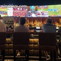A group of people sit at a bar with their backs to the camera, watching multiple sports games displayed on large screens. The bar counter is lined with various bottles of liquor, and the atmosphere suggests a casual and relaxed social setting.