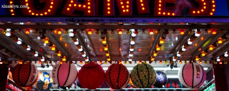 A brightly lit carnival game stall with a large 'GAMES' sign at the top surrounded by red and yellow lights. Various sports balls, including basketballs and soccer balls, are hanging from the ceiling and neatly arranged on shelves below.