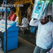 A street vendor stall displaying an array of lottery tickets clipped to a bright blue kiosk. A man in a red shirt leans on the counter, appearing thoughtful, while another man in a white shirt adjusts his hat and walks by. A variety of products hangs from strings above, and a colorful display fills the background with vibrant signs and packets. The scene suggests a busy, lively marketplace.